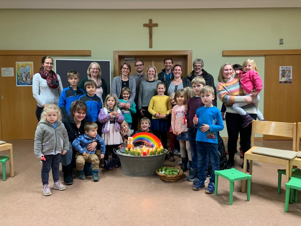 Um einen bunten Regenbogen stehen Kinder und Erwachsene zusammen. Es ist das Team und die Gäste der Familienkirche.